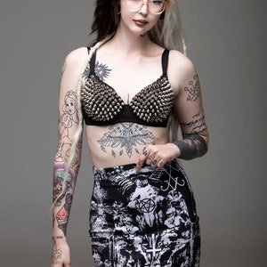 May include: A woman with black and blonde hair wearing a black bra with silver studs, a black and white printed mini skirt with the text "666, Lovis, Mayhem", and clear framed glasses. She has several tattoos on her arms and legs.