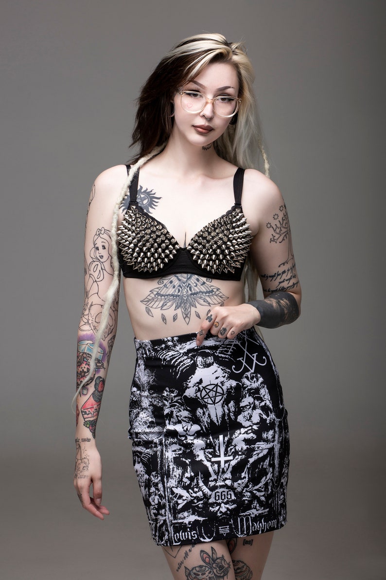 May include: A woman with black and blonde hair wearing a black bra with silver studs, a black and white printed mini skirt with the text "666, Lovis, Mayhem", and clear framed glasses. She has several tattoos on her arms and legs.