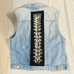 Op de afbeelding: Een lichtblauw denim vest met een zwart-witte ruggengraat grafische patch op de rug.