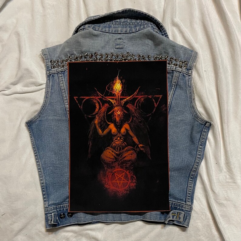 K&ouml;nnte beinhalten: Eine hellblaue Jeansweste mit einem gro&szlig;en Aufdruck auf der R&uuml;ckseite, der eine geh&ouml;rnte Figur mit einer Flamme &uuml;ber dem Kopf vor schwarzem Hintergrund zeigt. Die Weste ist mit silbernen Nieten am Kragen verziert.