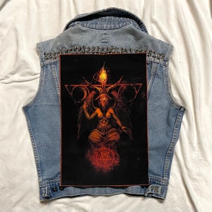 K&ouml;nnte beinhalten: Eine hellblaue Jeansweste mit einem gro&szlig;en, schwarzen Grafikaufn&auml;her, der eine geh&ouml;rnte Figur mit einer Flamme &uuml;ber dem Kopf zeigt. Die Weste ist mit silbernen Nieten am Kragen verziert.