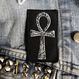 Puede incluir: Un parche termoadhesivo negro con un s&iacute;mbolo Ankh bordado en blanco. El Ankh es un s&iacute;mbolo jerogl&iacute;fico egipcio antiguo que representa la vida.