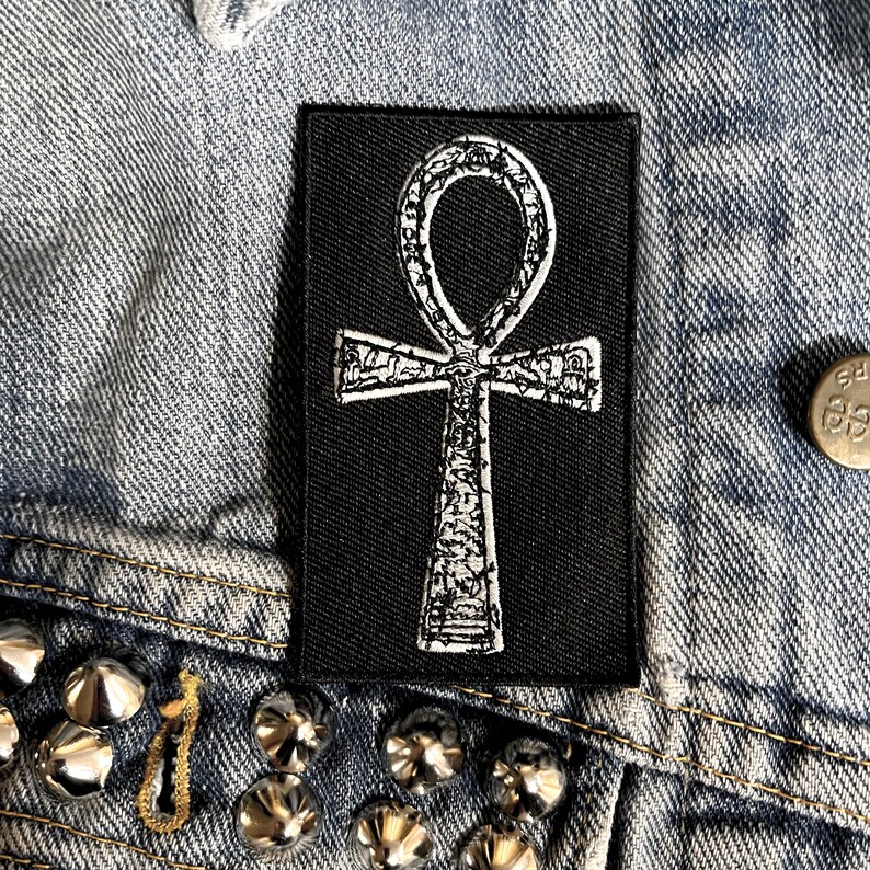 Puede incluir: Un parche termoadhesivo negro con un s&iacute;mbolo Ankh bordado en blanco. El Ankh es un s&iacute;mbolo jerogl&iacute;fico egipcio antiguo que representa la vida.
