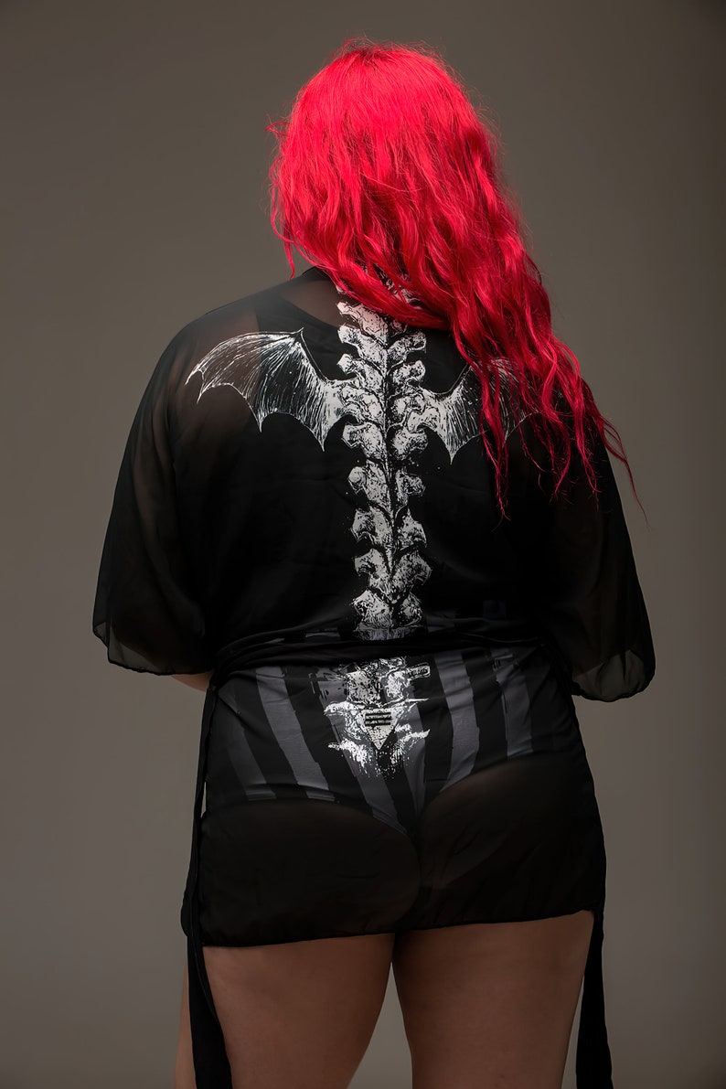 May include: Black sheer kimono-style top and shorts set with a white skeletal spine and bat wing design on the back. The shorts have a striped pattern. The model has bright red hair.