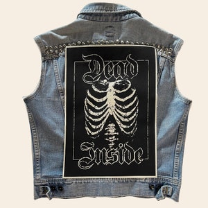 May include: A light blue denim vest with silver studs and a black patch on the back. The patch features a white skeleton ribcage with the words "Dead Inside" in a gothic font.