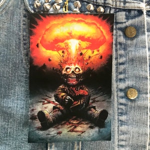 May include: A black and white patch featuring a cartoon character sitting in a bloody scene. The character has a skull head with glowing eyes and a mushroom cloud explosion above its head.