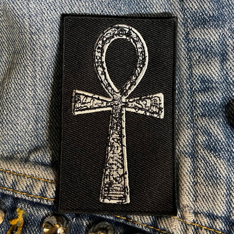 Puede incluir: Un parche de tela negro con un s&iacute;mbolo Ankh bordado en blanco. El Ankh es un s&iacute;mbolo de vida e inmortalidad en la religi&oacute;n egipcia antigua.