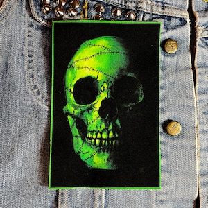 May include: Green skull with stitches on a black background. The skull is stitched together with yellow thread. The skull is a patch.