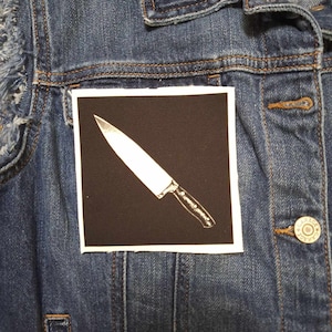 May include: A black and white iron-on patch with a white knife on a black background.