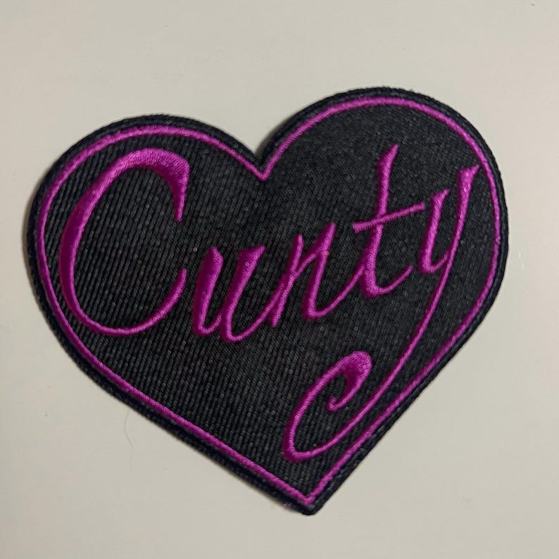 May include: A black heart-shaped patch with a magenta embroidered border and the word "CUNTY" in magenta script. The patch is made of a textured fabric.