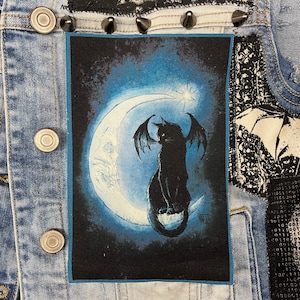 May include: Black cat with wings sitting on a crescent moon with a star in the background. The image is on a black fabric patch.