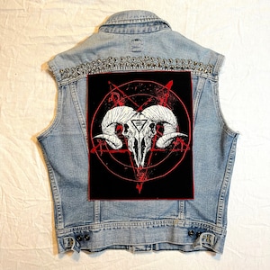 Op de afbeelding: Lichtblauwe spijkerbroekvest met zilveren studs op de schouders. Een grote zwarte patch op de rug toont een wit ram schedel ontwerp met een rode pentagram en andere rode grafische elementen. Het vest heeft een knoopsluiting.