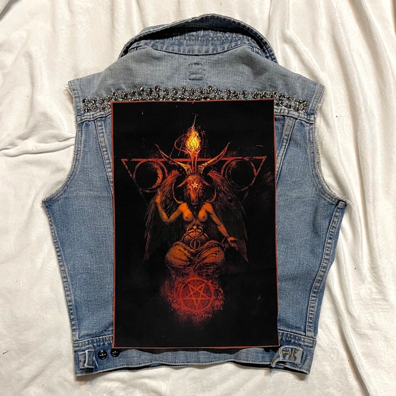 K&ouml;nnte beinhalten: Eine hellblaue Jeansweste mit einem gro&szlig;en, schwarzen Grafikaufn&auml;her, der eine geh&ouml;rnte Figur mit einer Flamme &uuml;ber dem Kopf zeigt. Die Weste ist mit silbernen Nieten am Kragen verziert.