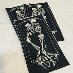 K&ouml;nnte beinhalten: Drei schwarze Stoffpatches mit wei&szlig;en gestickten Skeletten, die sich die H&auml;nde halten. Der Text "Ad Infinitum" befindet sich &uuml;ber den Skeletten. Der untere Patch hat ein Pentagramm mit der Zahl "23" darin.