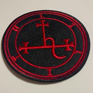 May include: A circular black patch with a red border and a red symbol in the center. The symbol appears to be a stylized cross with additional decorative elements. The patch is likely an emblem or insignia.