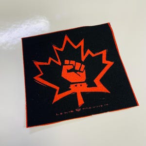 May include: A black fabric patch with a red maple leaf and a raised fist in the center. The text "Love & Mayhem" is visible at the bottom of the patch.
