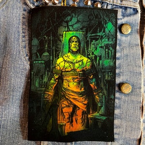 May include: A black and green embroidered patch depicting a Frankenstein-like figure with stitches and a belt, standing in a dark, green-lit room with various objects in the background.