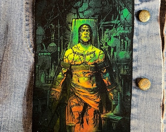 Puede incluir: Un parche bordado negro y verde con una figura de Frankenstein con puntadas y un cinturón, de pie en una habitación oscura iluminada en verde con varios objetos en el fondo.