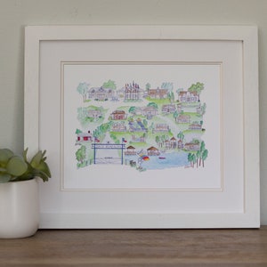 May include: A framed watercolor print of a town map, featuring various buildings, trees, and a lake. The print is in a white frame and has a white mat. The text "Sky Ranch" is visible on the print. A small potted plant sits to the left of the frame.