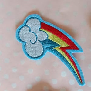 May include: Embroidered patch featuring a stylized rainbow and cloud design. The cloud is white with a light blue outline, and the rainbow displays red, yellow, and blue colors. The patch has a light blue border and is likely for clothing or accessories.