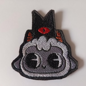 May include: Embroidered patch depicting a cartoon character with a black crown and a single red eye. The character has a grey face with large, black eyes and a black outline. The patch is designed for application on clothing or accessories.