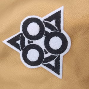 Pode incluir: Um patch bordado em branco e preto com um design geométrico com três círculos e triângulos. O patch está em uma superfície semelhante a couro bronzeado.