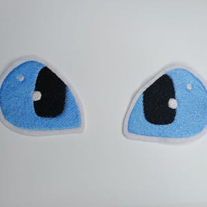 Könnte beinhalten: Zwei gestickte Aufnäher in Form blauer Augen. Die Augen haben eine schwarze Pupille, einen weißen Lichtreflex und eine hellblaue Iris. Die Aufnäher sind mit weißem Faden umrandet.
