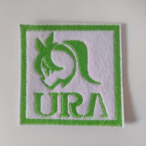 Op de afbeelding: Vierkante witte patch met een groene rand en een groen geborduurd ontwerp. Het ontwerp bevat een gestileerde vorm en de letters "URA" in het groen. De patch is waarschijnlijk voor kleding of accessoires.