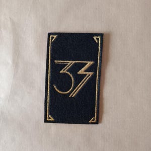 Könnte beinhalten: Ein rechteckiger, marineblauer Aufnäher mit goldener Stickerei. Das Design zeigt die Zahl "33" in einer stilisierten Schriftart, umrahmt von einem goldenen Rand. Der Aufnäher ist wahrscheinlich für Kleidung oder Accessoires.