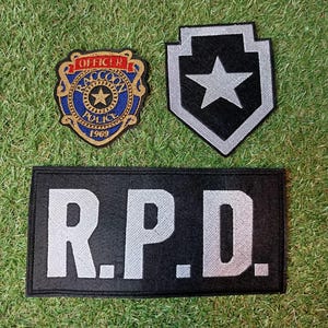Könnte beinhalten: Drei gestickte Aufnäher auf grünem Gras. Einer ist schildförmig mit einem weißen Stern auf schwarzem Hintergrund. Ein weiterer ist ein Abzeichen mit dem Text "OFFICER RACCOON POLICE 1969". Der dritte ist ein schwarzes Rechteck mit "R.P.D." in Weiß.