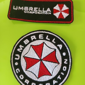 Pode incluir: Dois emblemas bordados com o logótipo da Umbrella Corporation. Um é um emblema retangular preto com texto branco e um ícone de guarda-chuva vermelho e branco. O outro é um emblema circular preto com texto branco e um ícone de guarda-chuva vermelho e branco.