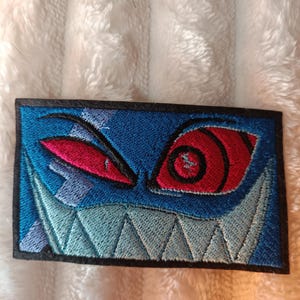 Könnte beinhalten: Gestickter Aufnäher mit einem stilisierten Monstergesicht mit roten Augen, scharfen Zähnen und blauem Hintergrund. Der Aufnäher hat einen schwarzen Rand und ist für Kleidung oder Accessoires geeignet.