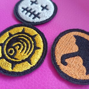 May include: Three embroidered patches with black borders. One is white with black crosses, another is gold and black with a spiral design, and the third is orange with a black umbrella silhouette. The patches are on a pink surface.