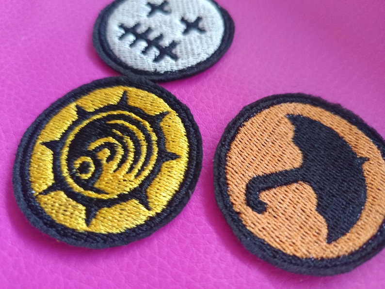 May include: Three embroidered patches with black borders. One is white with black crosses, another is gold and black with a spiral design, and the third is orange with a black umbrella silhouette. The patches are on a pink surface.