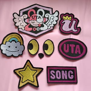 Op de afbeelding: Een verzameling van zeven geborduurde patches met diverse ontwerpen, waaronder een gevleugeld personage met de tekst UTA, een letter U met een kroon, een regenboogwolk, twee gele ogen, een ovale patch met UTA, een gouden ster en een rechthoek met SONG.