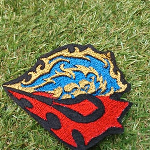 May include: An embroidered patch featuring a stylised design with a black border. The upper section displays a bright blue flame-like pattern outlined in gold thread, while the lower section consists of a bold red shape with black flame-like cutouts.