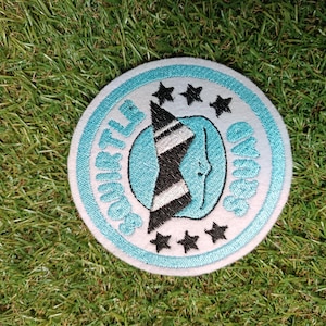 Op de afbeelding: Ronde geborduurde patch met de tekst "Squirtle Squad". Het ontwerp bevat een lichtblauwe rand en tekst, een zwart-witte afbeelding en zwarte sterren. De patch ligt op een groene ondergrond.