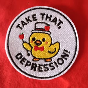 Könnte beinhalten: Gestickter Aufnäher mit einem gelben Cartoon-Küken, das einen Zylinder und eine Fliege trägt und einen roten Apfel an einem Stock hält. Der Text "TAKE THAT, DEPRESSION!" ist in einem kreisförmigen Design angeordnet.