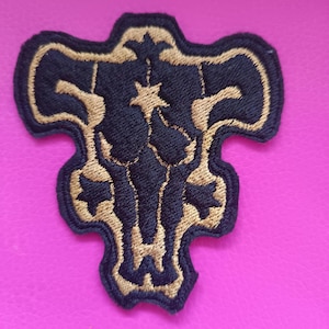 Può includere: Toppa ricamata nera e oro con un design a teschio di toro stilizzato. La toppa ha uno sfondo nero con cuciture color oro che delineano la forma e i dettagli. La toppa è su una superficie rosa.