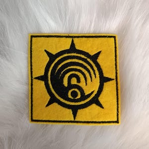 Può includere: Una toppa quadrata gialla con un emblema nero. L'emblema presenta un design circolare con una spirale e una stella a sei punte sul bordo. La toppa è probabilmente un articolo ricamato o in feltro.