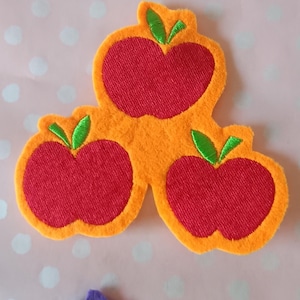 Könnte beinhalten: Drei Apfelformen aus Filz in Rot und Orange, mit grünen Blattakzenten. Die Äpfel sind zusammengefasst, mit einer genähten Textur. Der Hintergrund ist eine rosa Oberfläche mit weißen Punkten. Das Bild ist ein Bastelartikel.