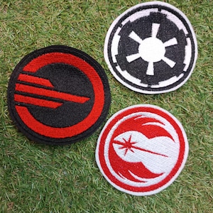 Può includere: Tre toppe ricamate circolari. Una toppa presenta un design rosso e nero con linee orizzontali. Un'altra è un emblema circolare bianco e nero con una stella centrale. La terza toppa è bianca con un design grafico circolare rosso.
