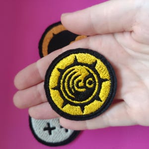 May include: Embroidered patches with a circular design. The primary patch is yellow and black, featuring a stylized emblem. Additional patches in the background include a black and orange design and a white patch with a black cross.