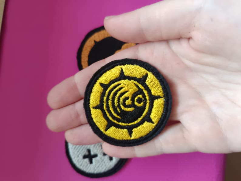 May include: Embroidered patches with a circular design. The primary patch is yellow and black, featuring a stylized emblem. Additional patches in the background include a black and orange design and a white patch with a black cross.