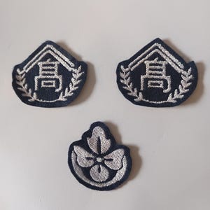 May include: Three embroidered patches. Two are navy blue with white text and a laurel wreath design. The third patch is a navy blue floral design with white embroidery. The patches are likely for clothing.