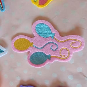 Könnte beinhalten: Rosa Filz-Applikation mit einem Design aus zwei blauen und einem gelben Ballon mit goldenen Schnurdetails. Andere farbige Filz-Applikationen umgeben es. Ideal für Bastel- und Verschönerungsprojekte.