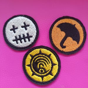 Könnte beinhalten: Drei runde, gestickte Aufnäher. Einer hat ein gesticktes Gesichtsdesign auf weißem Hintergrund. Ein anderer zeigt einen schwarzen Regenschirm auf orangefarbenem Hintergrund. Der dritte hat ein gelb-schwarzes Design.