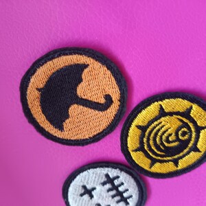 May include: Three embroidered patches with black and orange designs on a pink background. One patch features a black umbrella on an orange circle. Another has a yellow sunburst design. The third patch has a white background with black stitches.