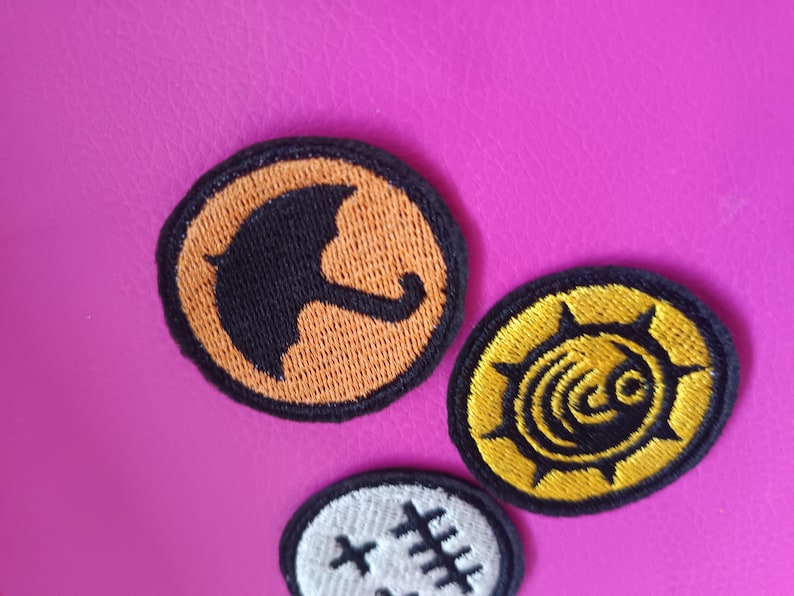 May include: Three embroidered patches with black and orange designs on a pink background. One patch features a black umbrella on an orange circle. Another has a yellow sunburst design. The third patch has a white background with black stitches.