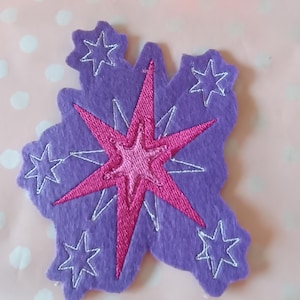 Könnte beinhalten: Lila Filz-Applikation mit einem pinkfarbenen Sternenmuster und weißen gestickten Sternen. Dieses Bastel-Element könnte zur Dekoration von Kleidung oder Accessoires verwendet werden. Auf rosa Pünktchen-Hintergrund.
